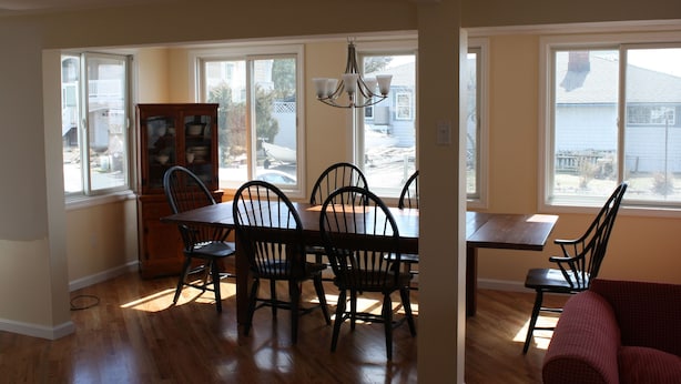 A dining area with a wooden table and chairs, a chandelier, and large windows with a view of a house and trees. - 6 Beds, 4 Baths w Elevator LBI - Now Booking 2027 limited 2026 dates still avail (Beach Haven)