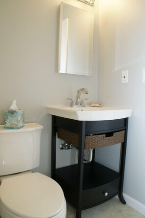 A bathroom with a white toilet, a sink with a mirror above it, and a basket under the sink. - Carefree Days at The Beach (Watsonville)