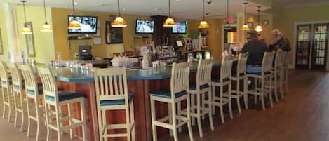 A bar area with multiple bar stools, a counter with a cash register, and overhead televisions displaying content.