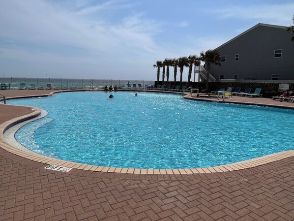 Une piscine entourée d’un sol en briques, bordée de palmiers et de chaises longues, avec un bâtiment visible au loin.