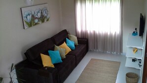 A living room with a dark brown sofa, colorful cushions, a large window with sheer curtains, a ceiling fan, a mounted TV, and a small plant. - Casa Verde Paraty (Paraty)