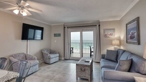 A spacious living room with a ceiling fan, a flat-screen TV, a sofa, a coffee table, and a view of the ocean through a sliding glass door. - Seacrest 405 1 Bedroom Condo by RedAwning (Gulf Shores)