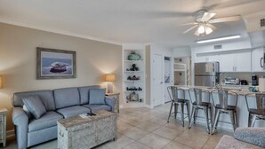 A living room with a grey sofa, a coffee table, and a painting on the wall. There is a kitchen area with a refrigerator, stove, and bar stools. - Seacrest 405 1 Bedroom Condo by RedAwning (Gulf Shores)