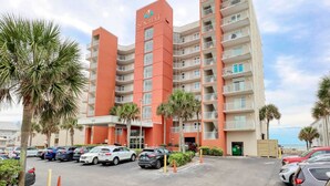 A Seacrest hotel with a red facade, multiple balconies, and a parking lot with several cars. - Seacrest 405 1 Bedroom Condo by RedAwning (Gulf Shores)