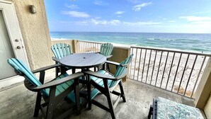 A balcony with a round table and four chairs, overlooking a beach with the ocean and a clear sky.