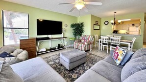 A living room with a TV, sofa, ottoman, and a dining area with chairs and a table. - Sea Oats H102 2 Bedroom Condo by RedAwning (Gulf Shores)