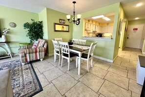 A dining area with a table and chairs, a sofa, a chandelier, and a kitchen in the background. - Sea Oats H102 2 Bedroom Condo by RedAwning (Gulf Shores)