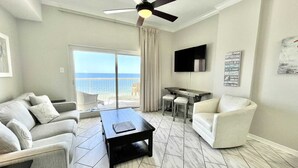 A modern living room with a ceiling fan, a flat-screen TV, a dining area, and a view of the ocean. - Royal Palms 1306 1 Bedroom Condo by RedAwning (Gulf Shores)