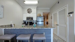 A kitchen with a central island, bar stools, a microwave, a refrigerator, and a decorative wall piece. - Royal Palms 1306 1 Bedroom Condo by RedAwning (Gulf Shores)