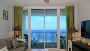 A living room with a sofa, a coffee table, and a TV. There is a balcony with a view of the ocean. - Lighthouse 1017 1 Bedroom Condo by RedAwning (Gulf Shores)
