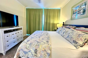 A hotel room with a large bed, a TV on a dresser, a painting on the wall, and green curtains.