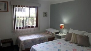 A bedroom with two beds, a nightstand, a window with bars, and a wall-mounted lamp. - Casa Condomínio Geribá (Búzios)