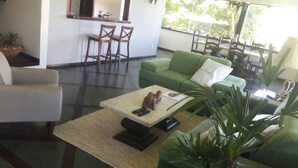 Una sala de estar con un sofá verde, un sillón blanco y una mesa de centro con mandos a distancia. Se aprecia una pequeña mesa con sillas al fondo.