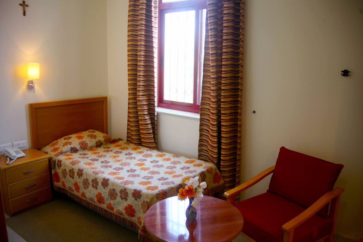 A hotel room with a bed, a chair, a bedside table, a lamp, and a window with curtains.