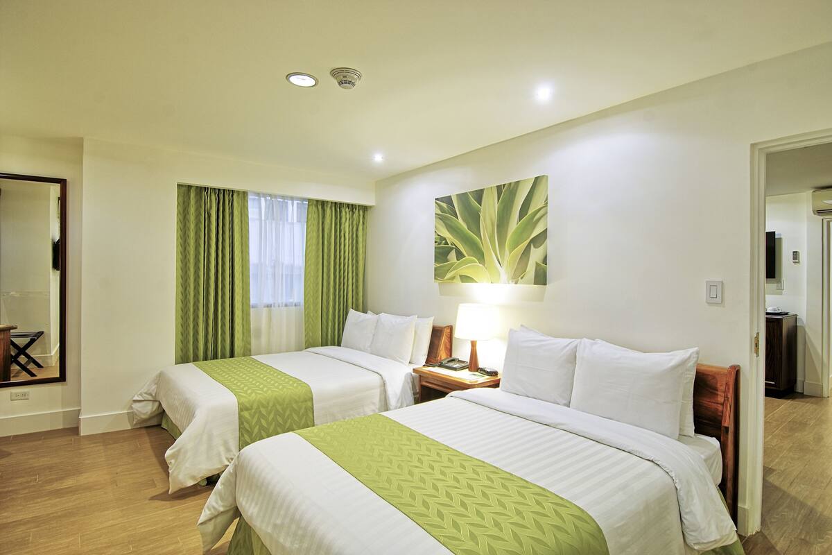 A hotel room with two beds, a nightstand with a lamp, a large window with curtains, and a mirror on the wall.