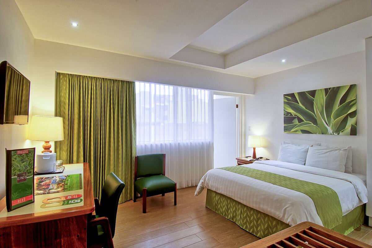 A hotel room with a bed, a desk with a lamp, a chair, a window with curtains, and a wall-mounted artwork.