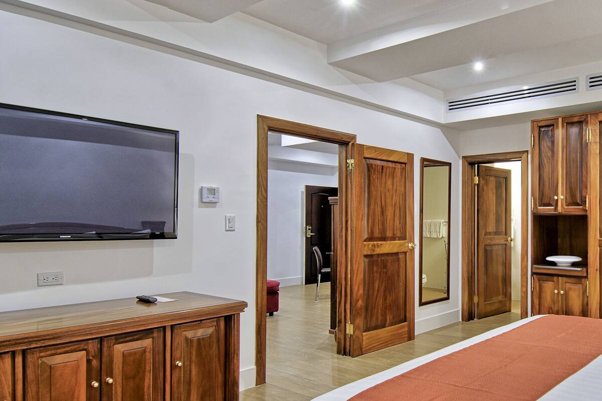A hotel room with a large bed, wooden wardrobe, a flat-screen TV mounted on the wall, and a bathroom visible through an open door.