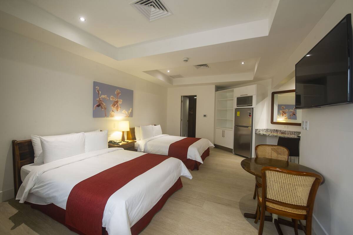 A hotel room with two beds, a dining table with chairs, a TV, and a kitchenette.