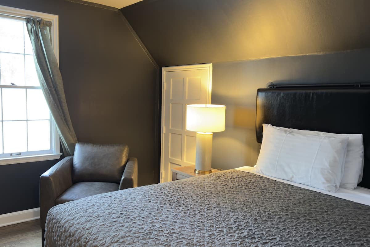 Een slaapkamer met een bed, een stoel, een nachtkastje met lamp, een raam met gordijnen en een deur.