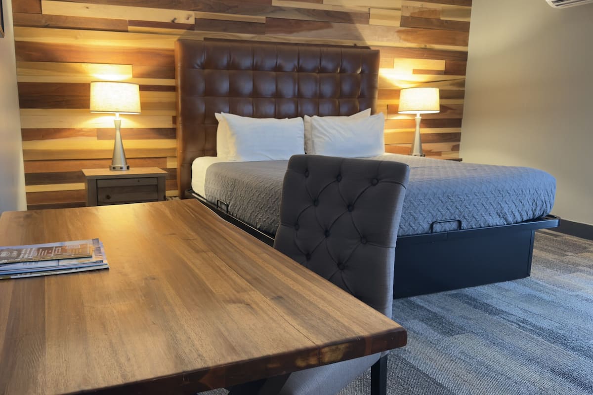 Een hotelkamer met een houten hoofdeinde, een bed met een grijze deken, twee bedlampjes, een nachtkastje van hout, en een houten tafel met een tijdschrift.