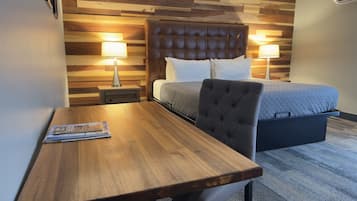 Habitación de hotel con cabecera de madera, cama con colcha gris, dos lámparas de noche, una mesita de noche de madera y una mesa de madera con una revista.
