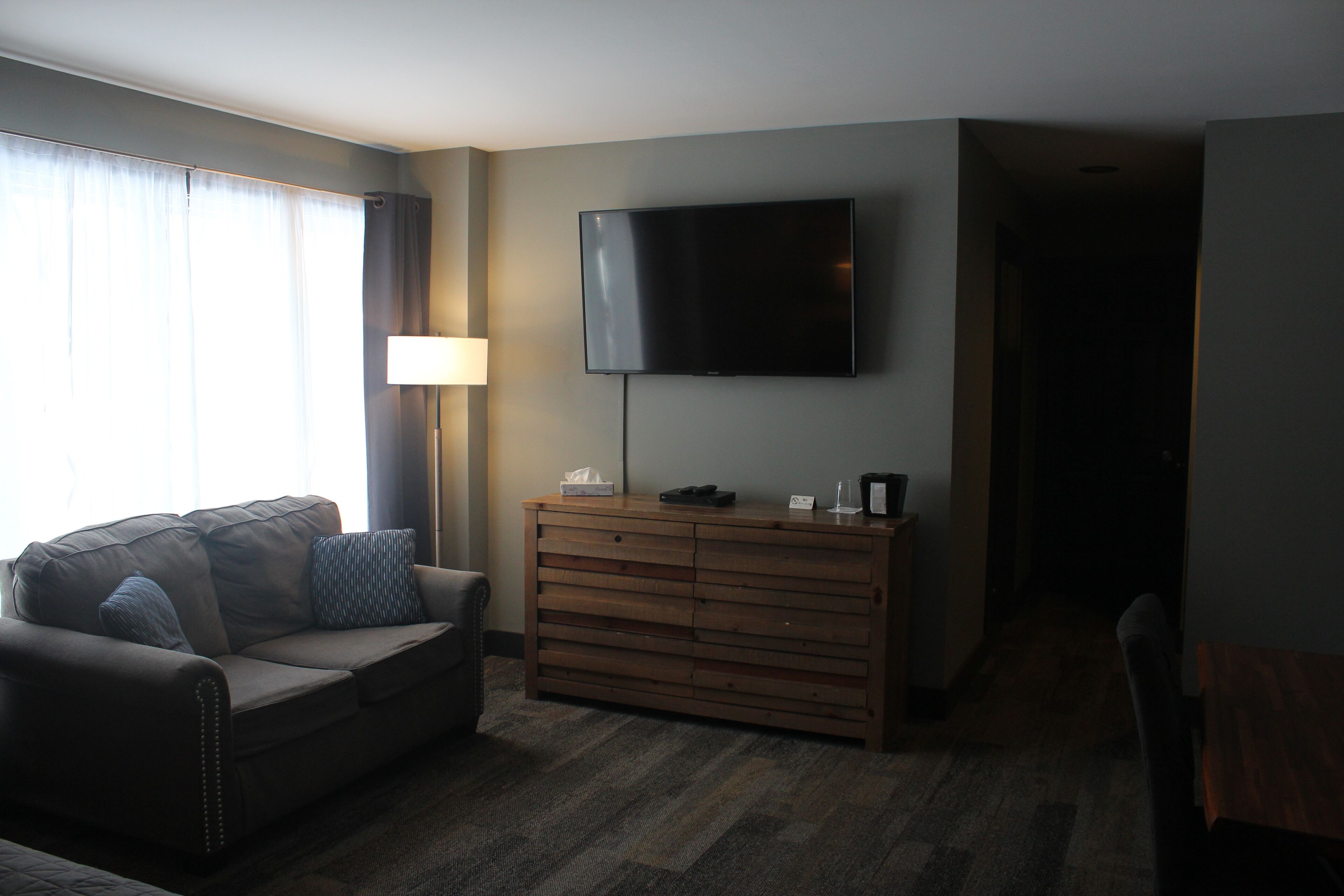 A hotel room with a sofa, a wooden dresser, a flat-screen TV mounted on the wall, and a lamp.