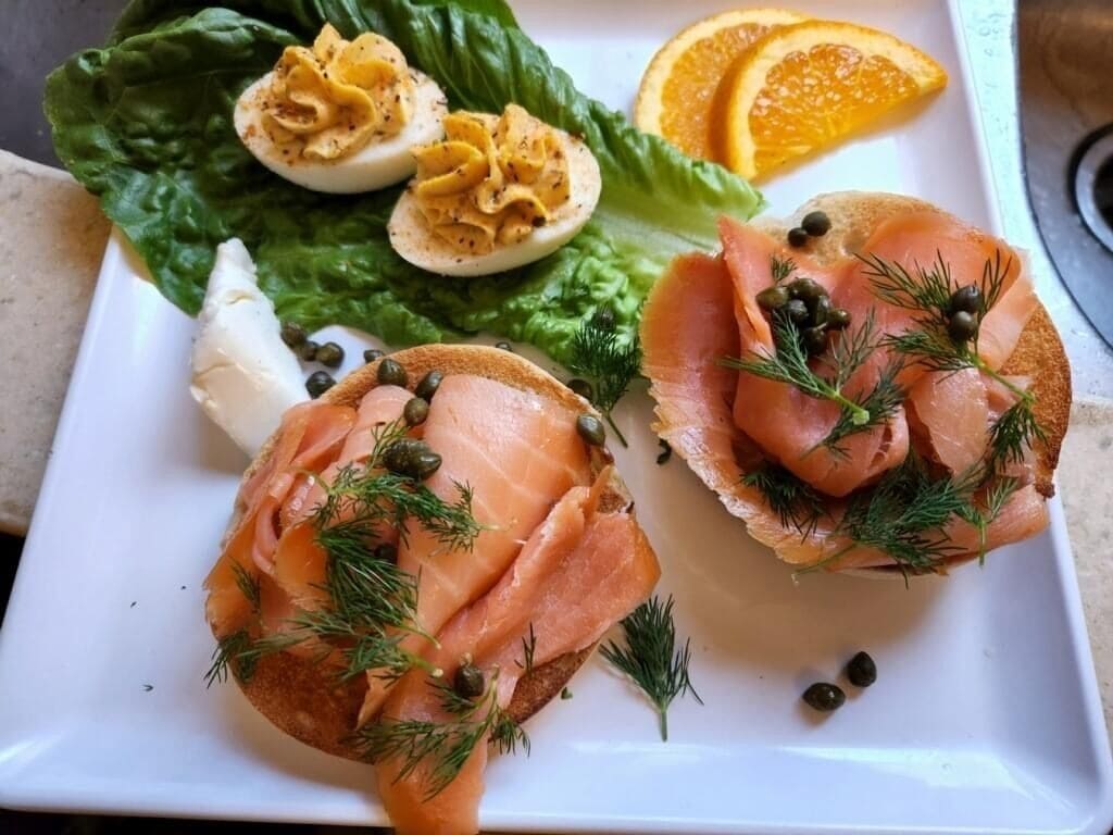 A plate with two open-faced sandwiches topped with smoked salmon and capers, a boiled egg, and a side of orange slices.