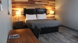 Een slaapkamer met een houten hoofdbord, twee nachtkastjes met lampen, een bed met een grijze quilt en een houten bureau met een tijdschrift.