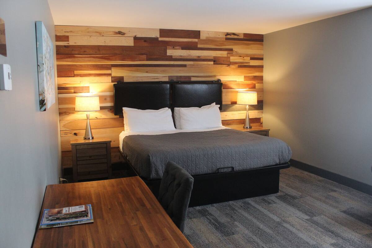 Een slaapkamer met een houten hoofdbord, twee nachtkastjes met lampen, een bed met een grijze quilt en een houten bureau met een tijdschrift.