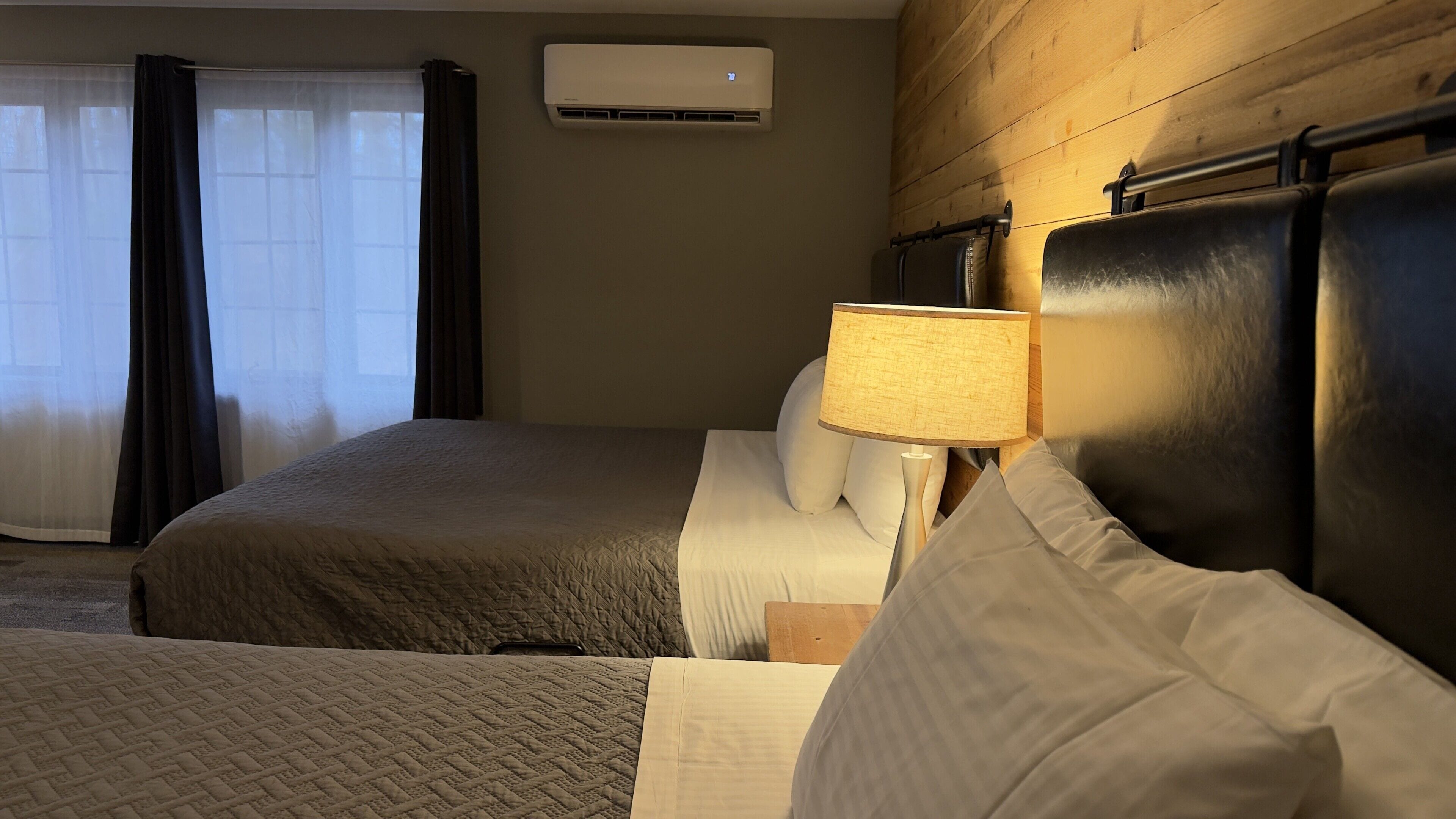 A hotel room with a bed, a bedside lamp, a window with curtains, and an air conditioning unit.