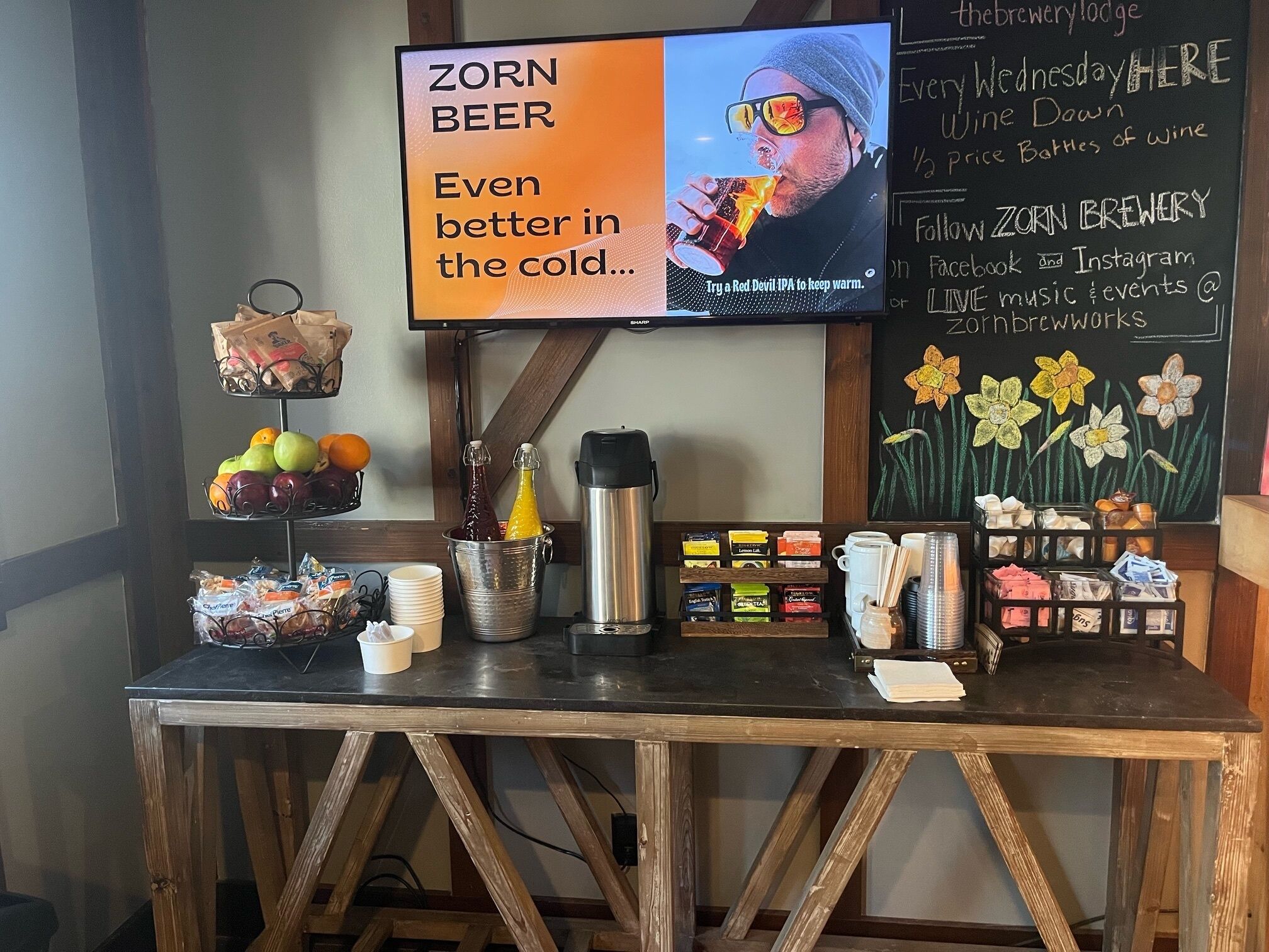 A beverage station with a sign promoting Zorn Beer, a selection of fruits, and a chalkboard with event information.