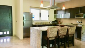 A modern kitchen with a central island, bar stools, stainless steel appliances, and pendant lighting. - Mamitas Village by Casago (Playa del Carmen)
