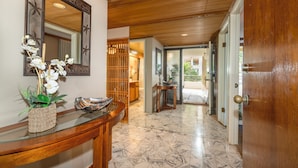Um corredor com teto de madeira, uma mesa de console de vidro com uma planta, um espelho e chão de azulejos.