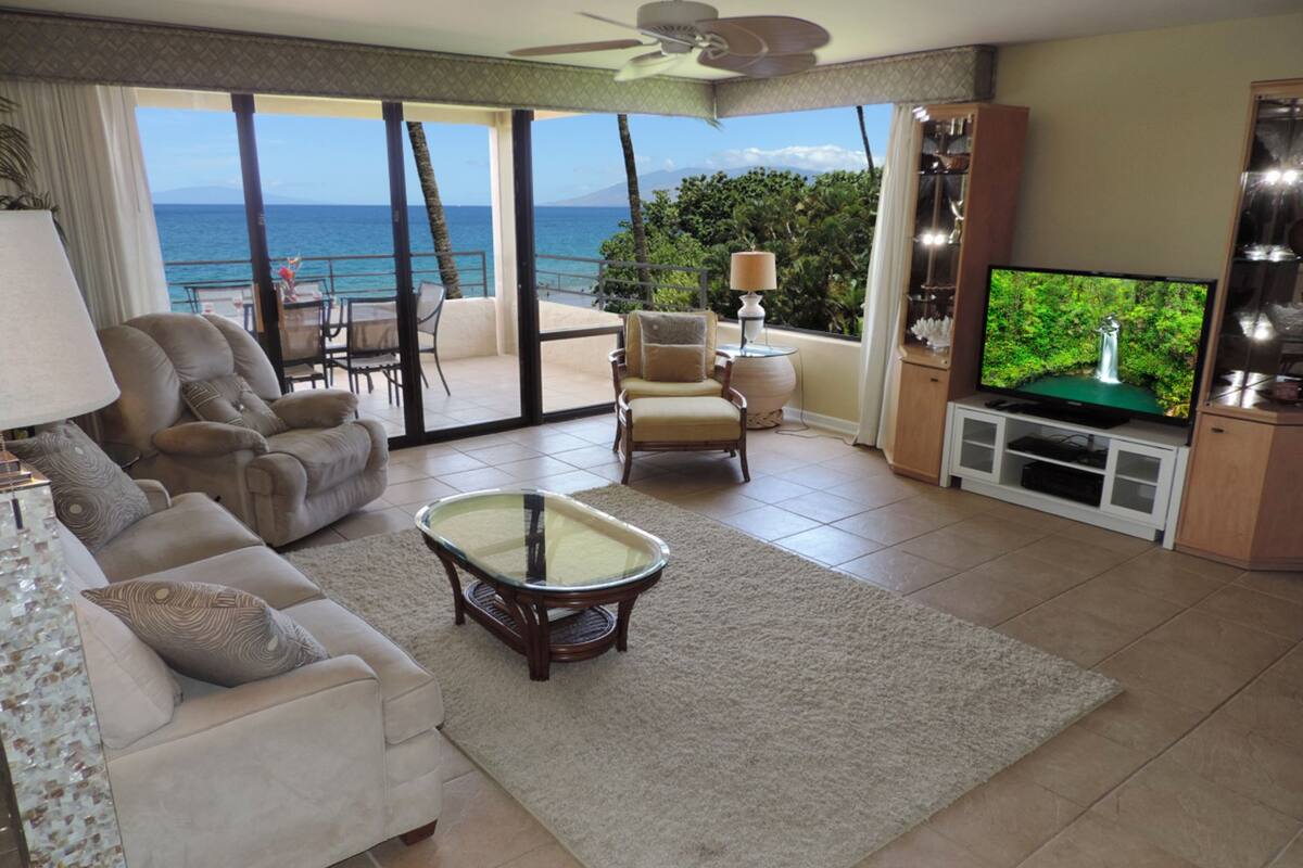 A living room with a large TV, comfortable seating, and a view of the ocean.