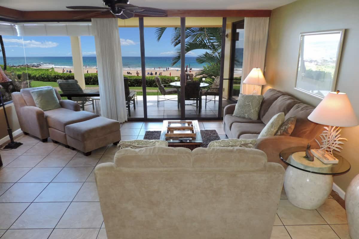 A living room with a view of the beach, a ceiling fan, and a glass coffee table.