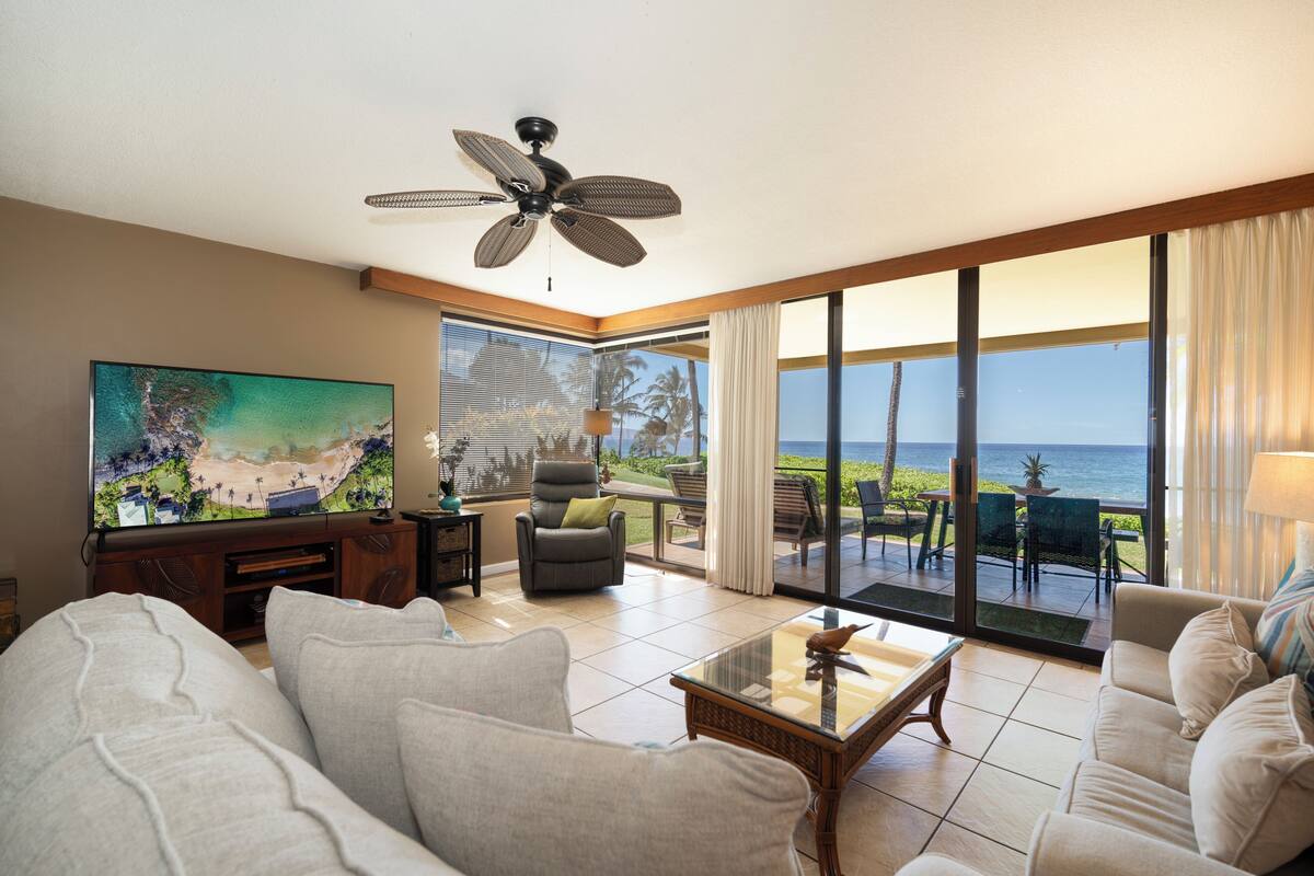 A spacious living room with a large TV, comfortable seating, a ceiling fan, and a view of the ocean.