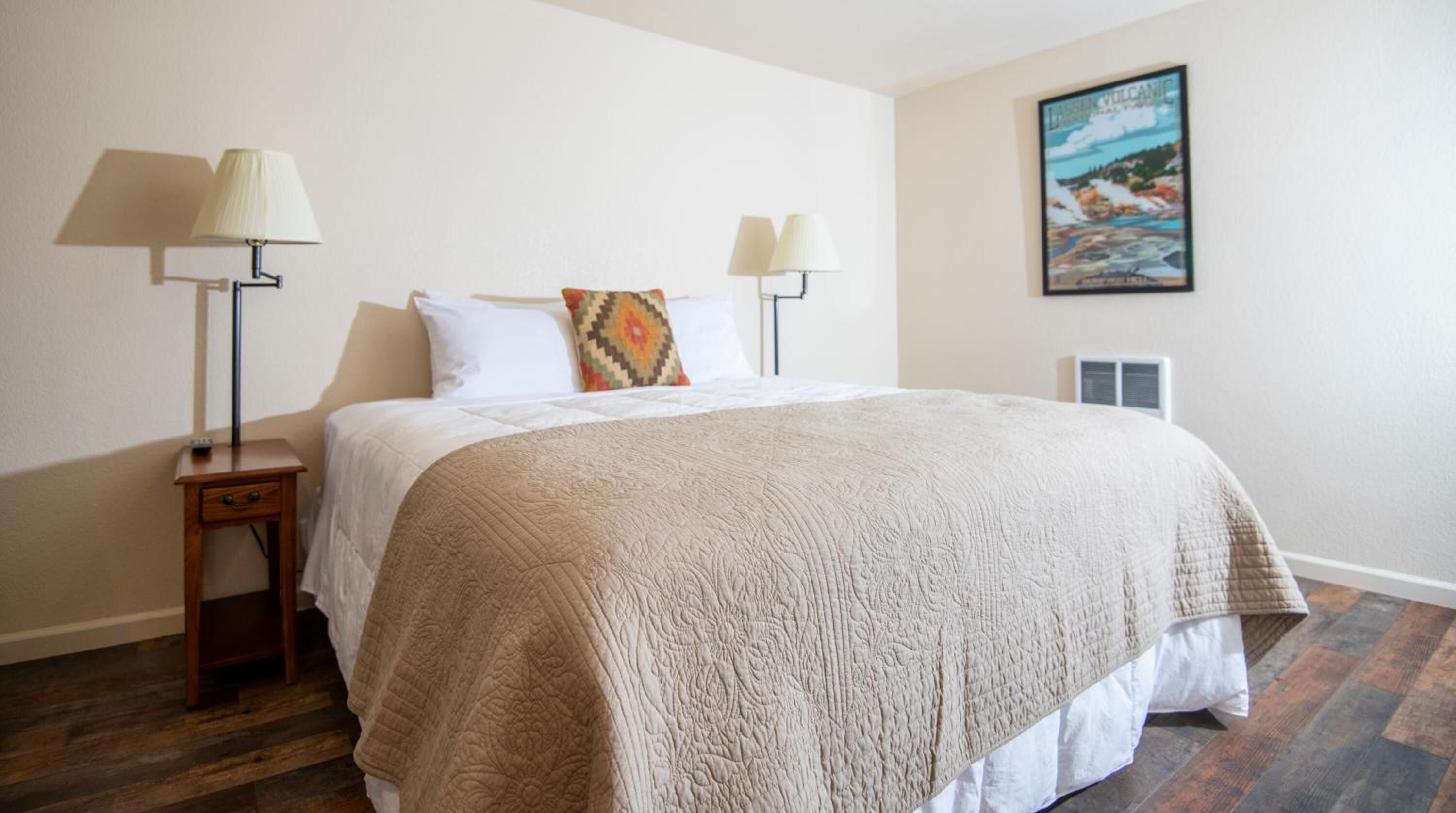 A neatly arranged bedroom with a large bed, two bedside lamps, a small bedside table, and a wall-mounted air conditioning unit.