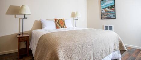 A neatly arranged bedroom with a large bed, two bedside lamps, a small bedside table, and a wall-mounted air conditioning unit.