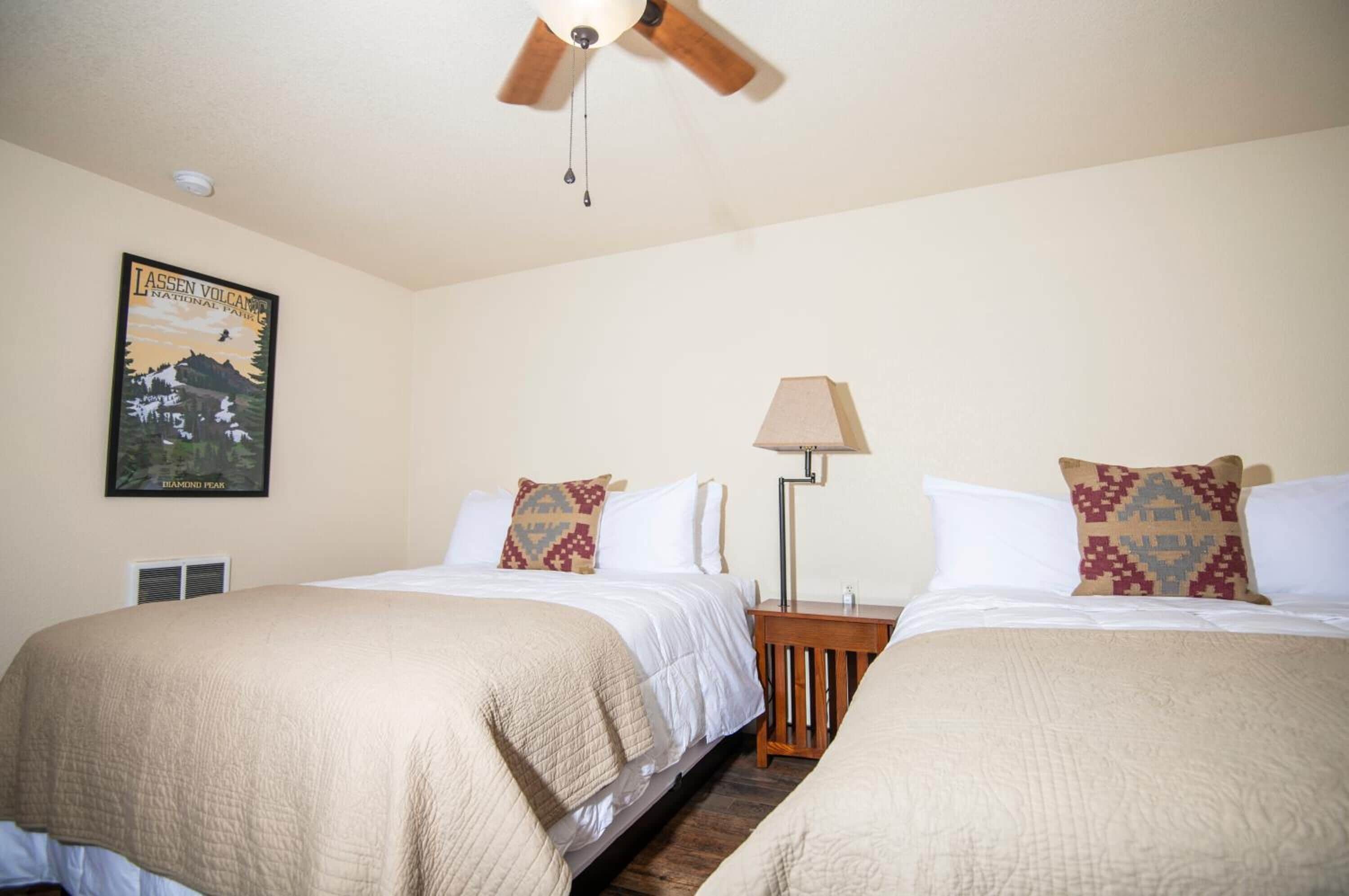 A hotel room with two beds, a ceiling fan, a lamp, and a poster on the wall.