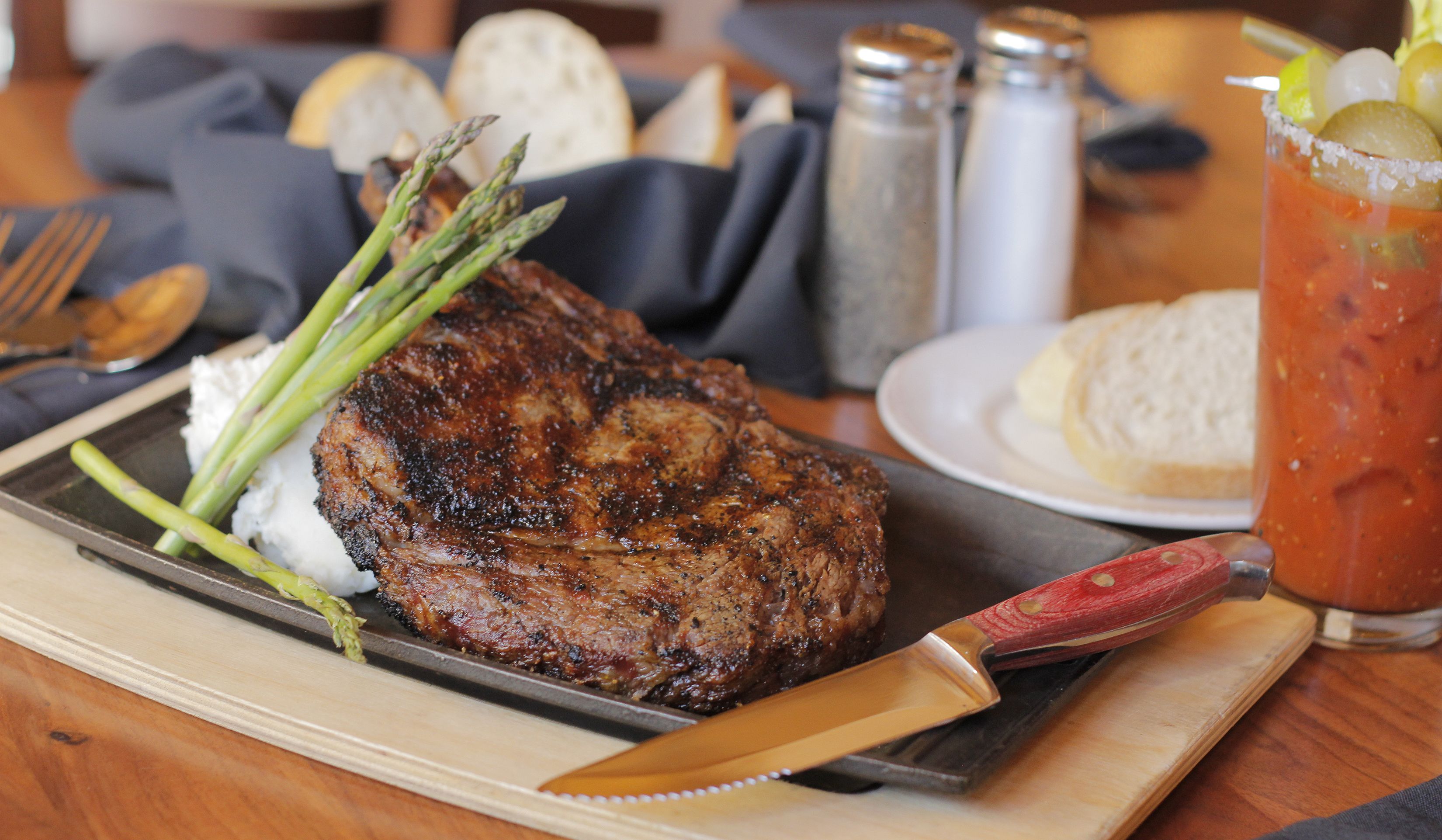 Un steak accompagné d’asperges, un couteau sur une assiette, un verre rempli d’une boisson, et des salières et poivrières sur une table.