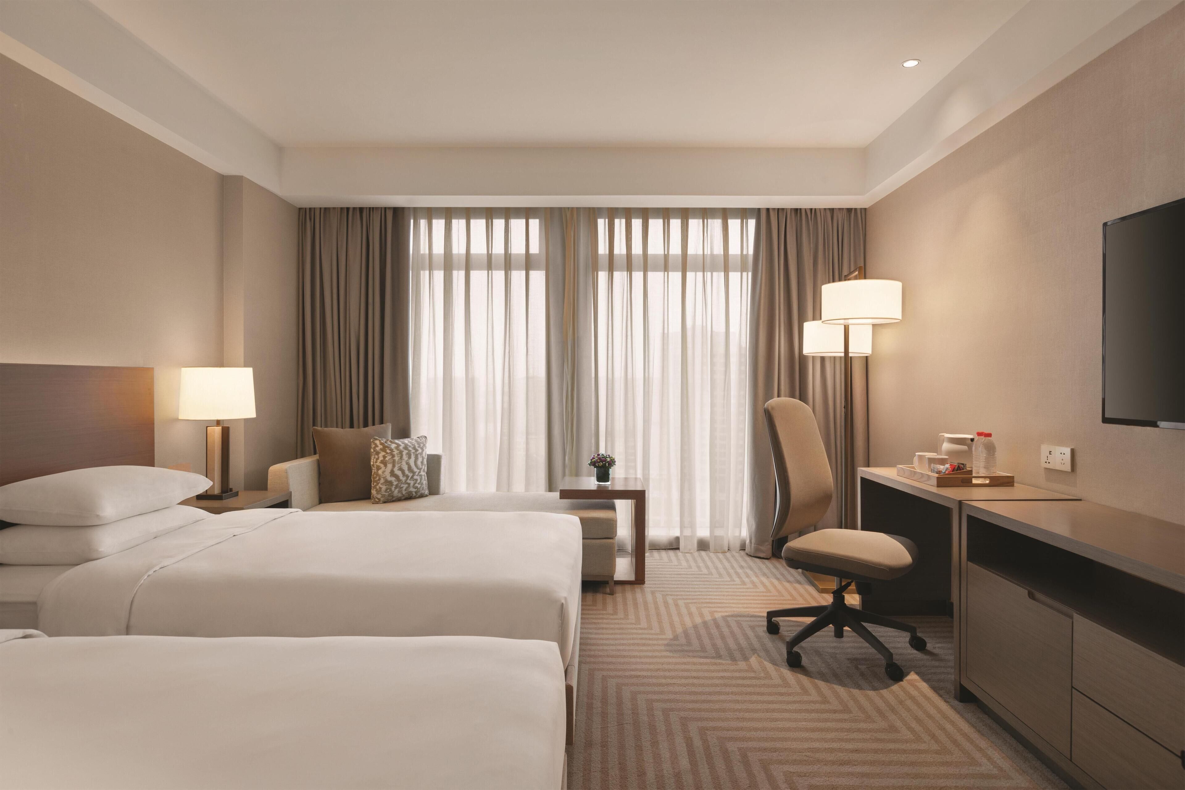 Une chambre d’hôtel avec deux lits, un bureau, une chaise, une télévision et une fenêtre avec des rideaux.