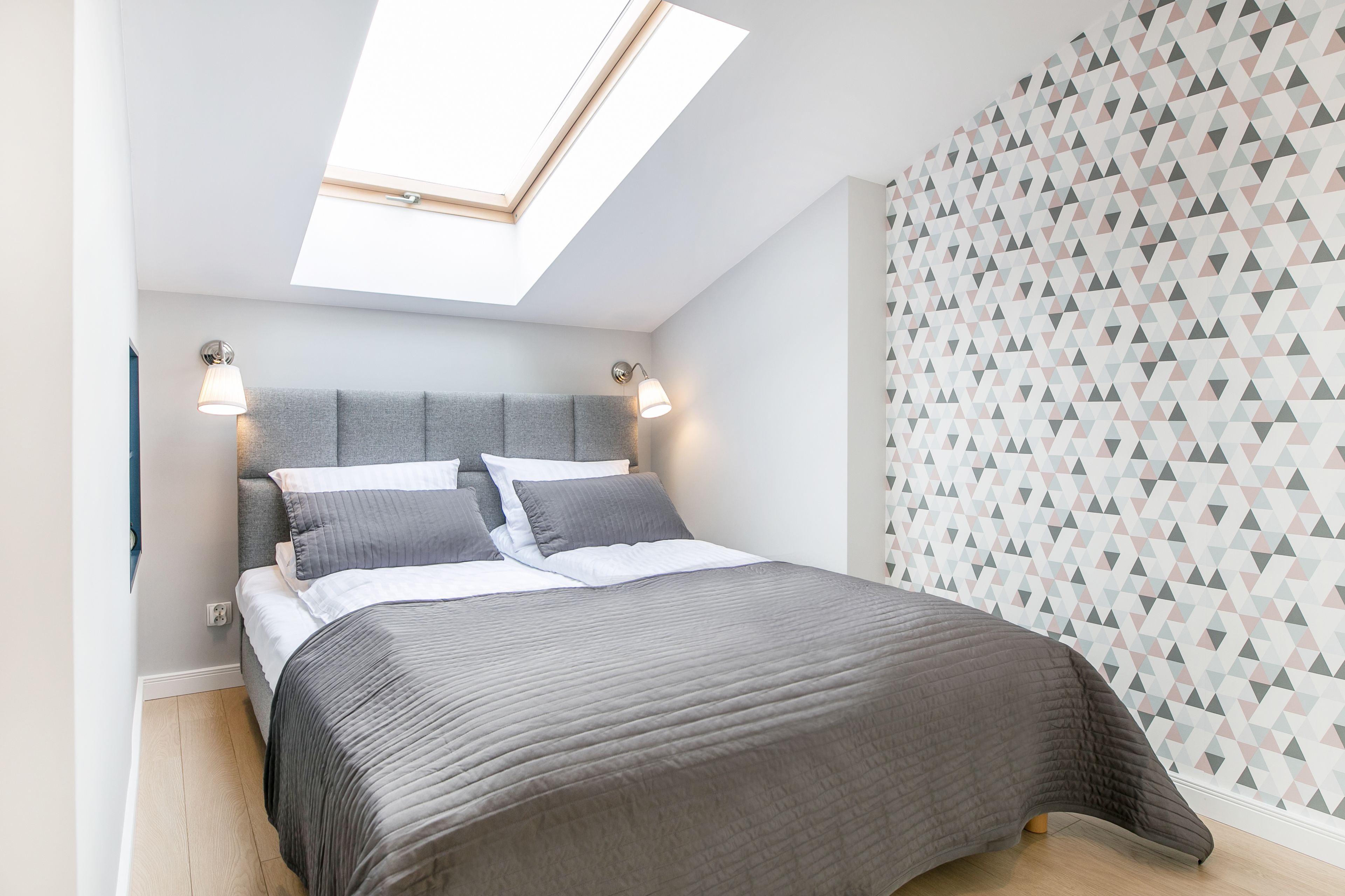 Een slaapkamer met dakraam, een bed met grijze beddengoed en een behang met patroon.