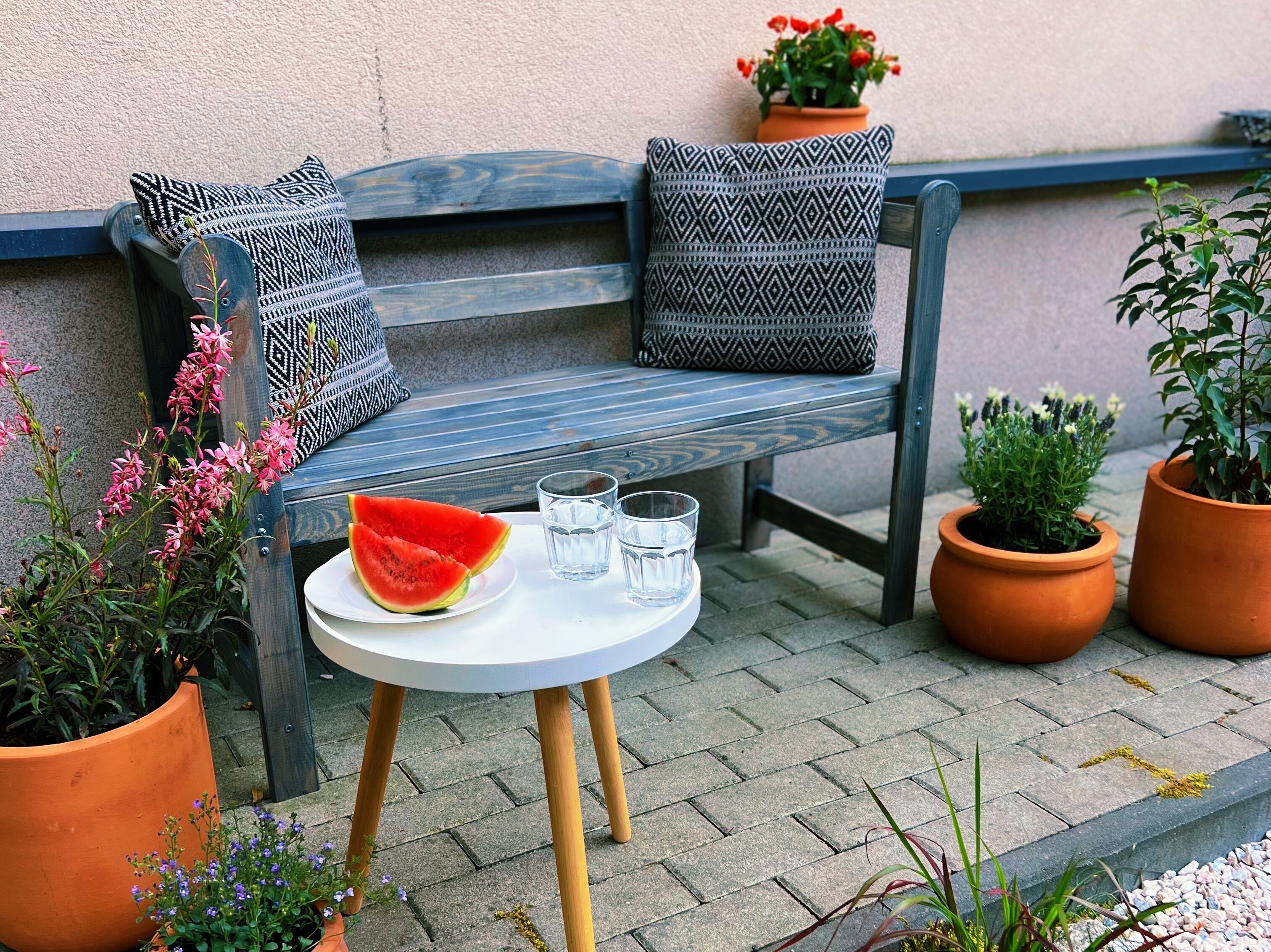 A wooden bench with patterned cushions, a small table with a watermelon slice and glasses, and potted plants around.
