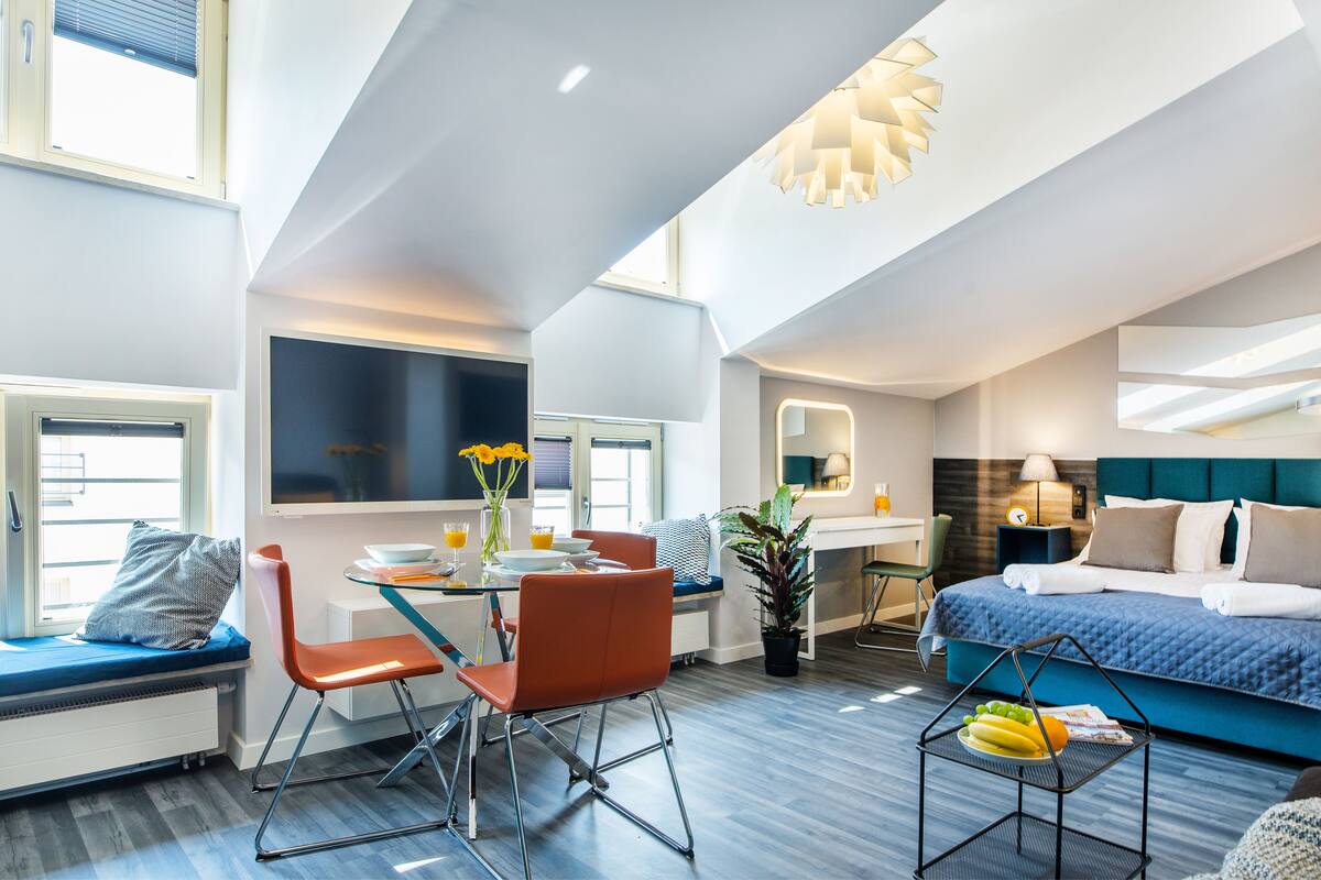 A modern hotel room with a bed, a dining area, and a seating area with a window.