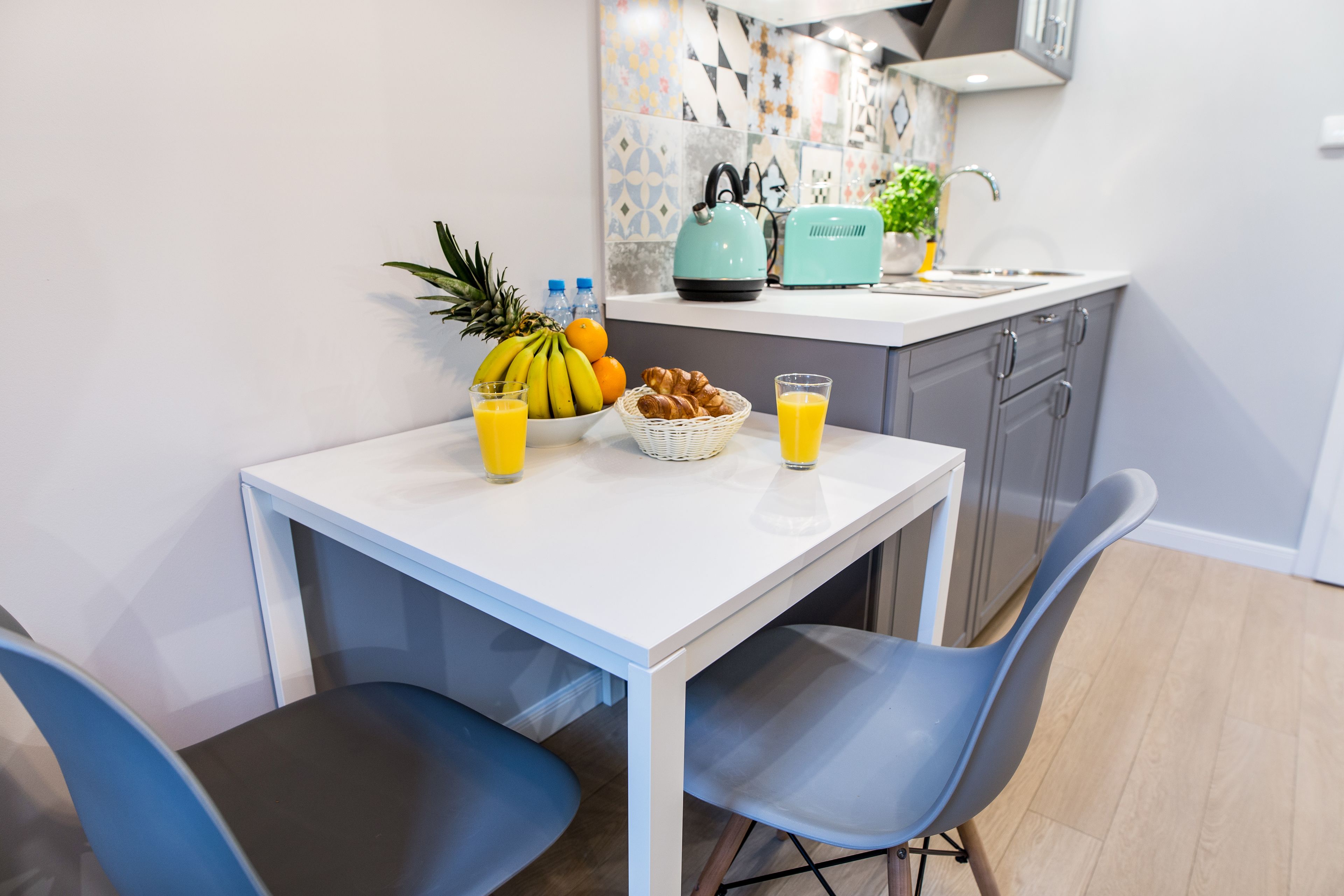 A modern kitchen with a white table, blue chairs, a fruit basket, and a toaster.