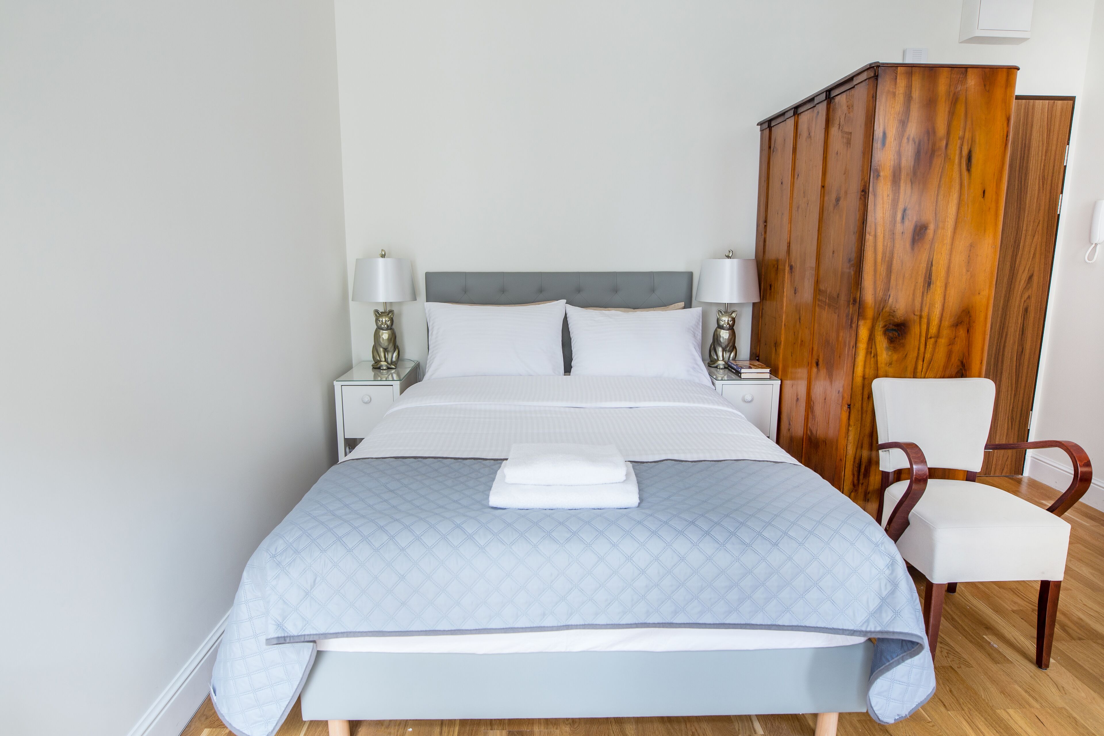 A neatly made bed with a quilted blue cover, two bedside lamps, a wooden wardrobe, and a white chair.