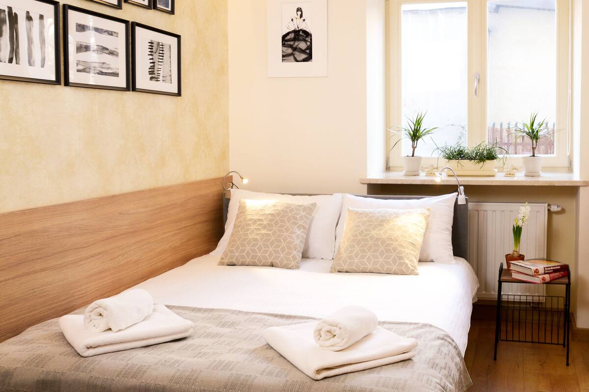 A neatly made bed with rolled towels, a bedside table with books and a plant, and a window with a view of buildings.