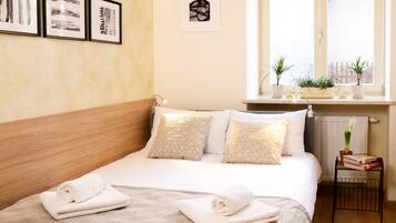 A neatly made bed with rolled towels, a bedside table with books and a plant, and a window with a view of buildings.