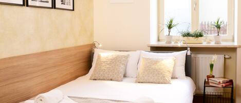 A neatly made bed with rolled towels, a bedside table with books and a plant, and a window with a view of buildings.