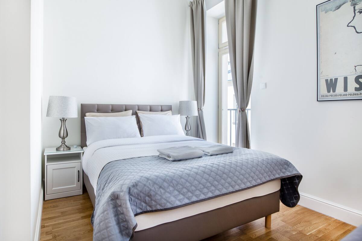 A neatly made bed with a grey quilt, two bedside lamps, and a small nightstand with a lamp.